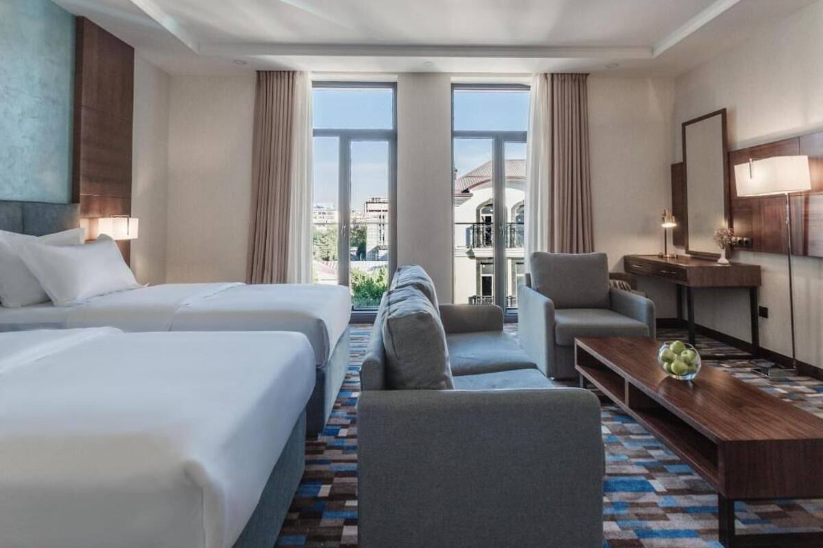 A hotel room with two beds, a sofa, a chair, a desk, and a large window offering a city view.