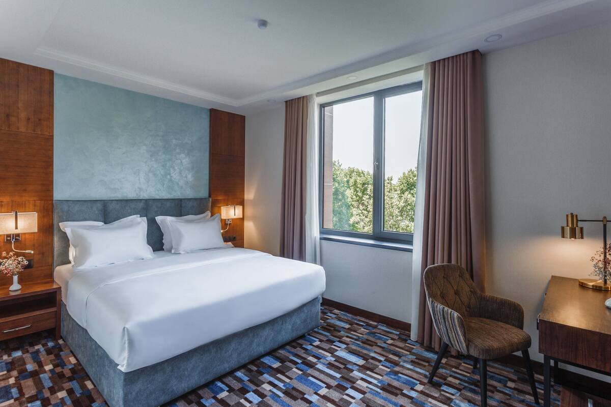 A hotel room with a large bed, a chair, a desk, and a window with curtains.