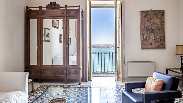 Una habitación con un amplio armario de madera, una mesa con un cuenco, un sofá, una silla, un cuadro y vistas al mar a través de una puerta abierta.