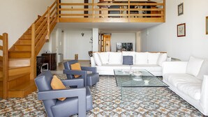 Amplia sala de estar con un sofá blanco, una mesa de centro de vidrio y un sillón azul con cojín. Una escalera de madera conduce a un nivel superior con una litera.