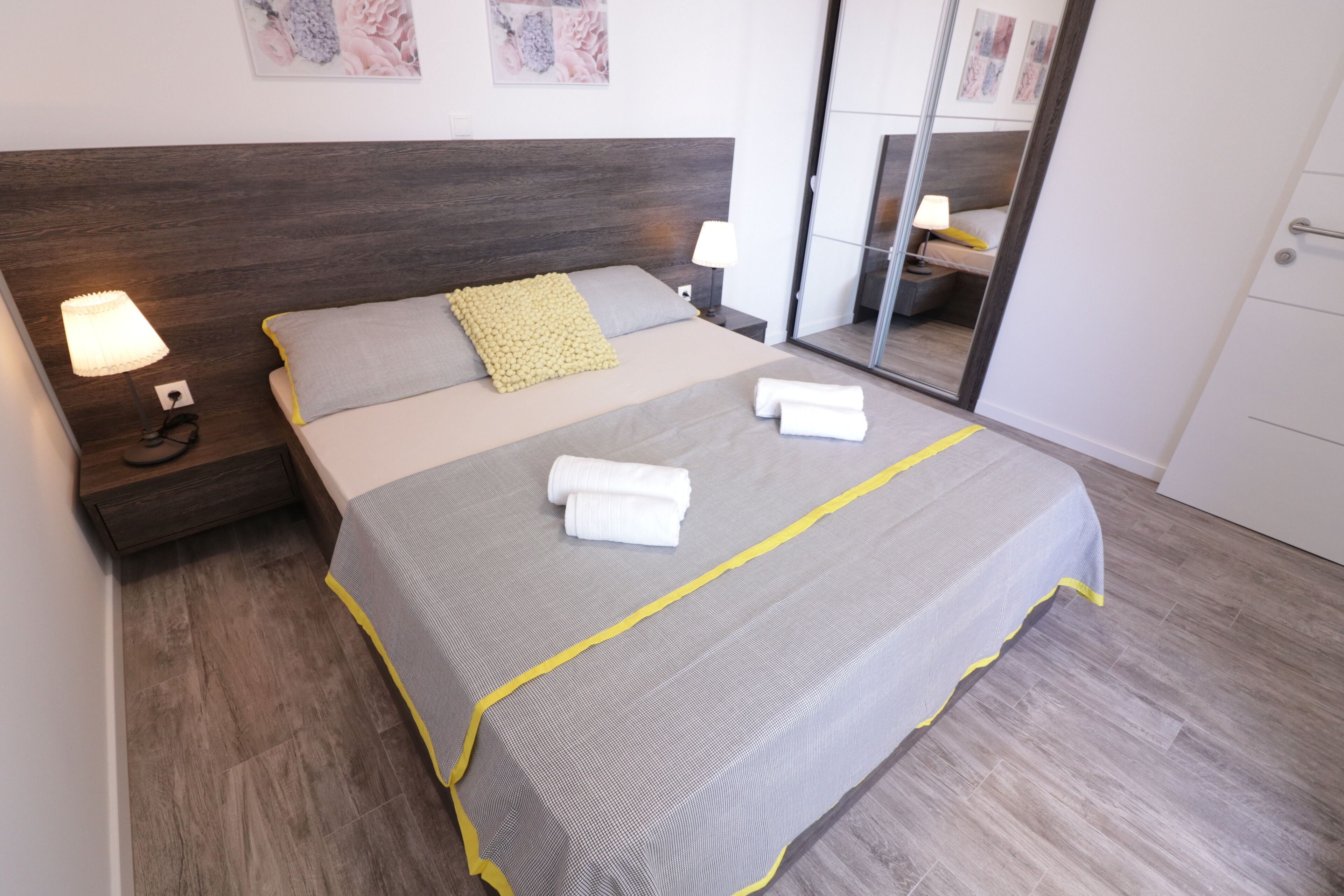 Un lit bien fait, recouvert d’une couvre-lit grise et jaune, avec deux oreillers et deux serviettes blanches pliées. Il y a une table de chevet avec une lampe et une prise électrique. La pièce est dotée d’un plancher en bois et d’un grand miroir sur le mur.