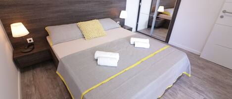 A neatly made bed with a grey and yellow bedspread, two pillows, and two folded white towels. There is a nightstand with a lamp and a power outlet. The room has wooden flooring and a large mirror on the wall.