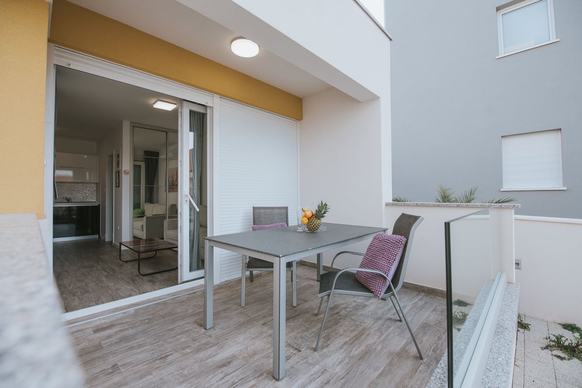 An outdoor balcony with a table and chairs, a sliding glass door leading to an interior space, and a small plant on the wall.