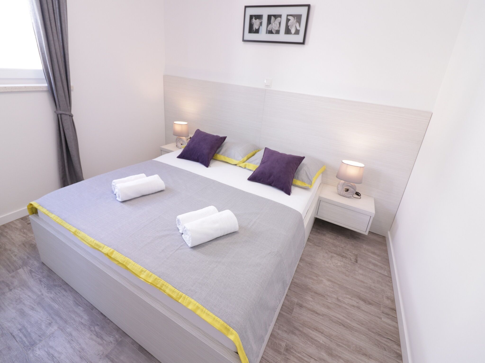 A neatly made bed with a grey and yellow bedspread, two purple pillows, and two white towels. There are two bedside lamps, a framed picture on the wall, and a window with curtains.