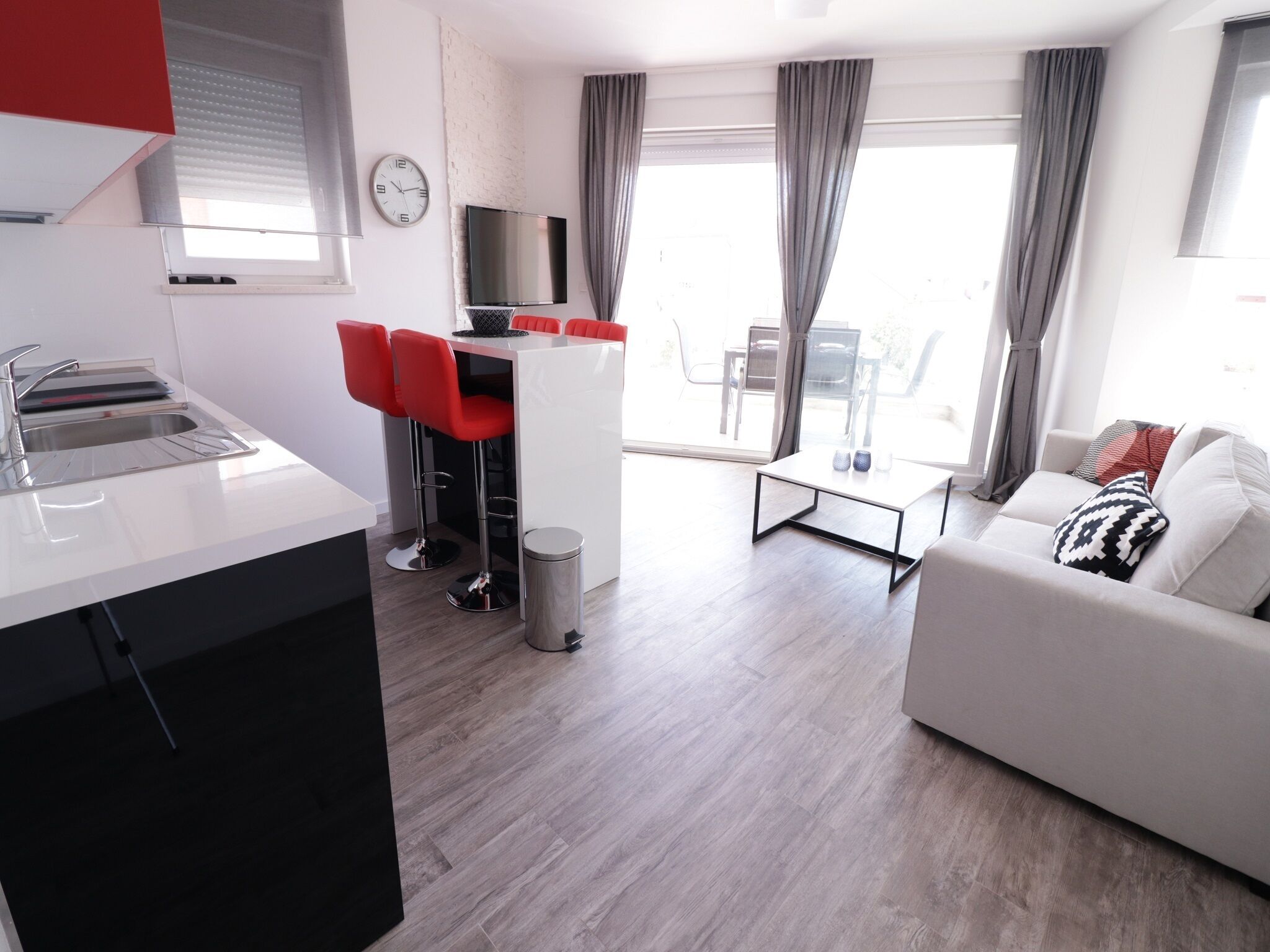 A modern apartment with a kitchen, dining area, and living room. It includes a white sofa, a dining table with red chairs, and a TV mounted on the wall.
