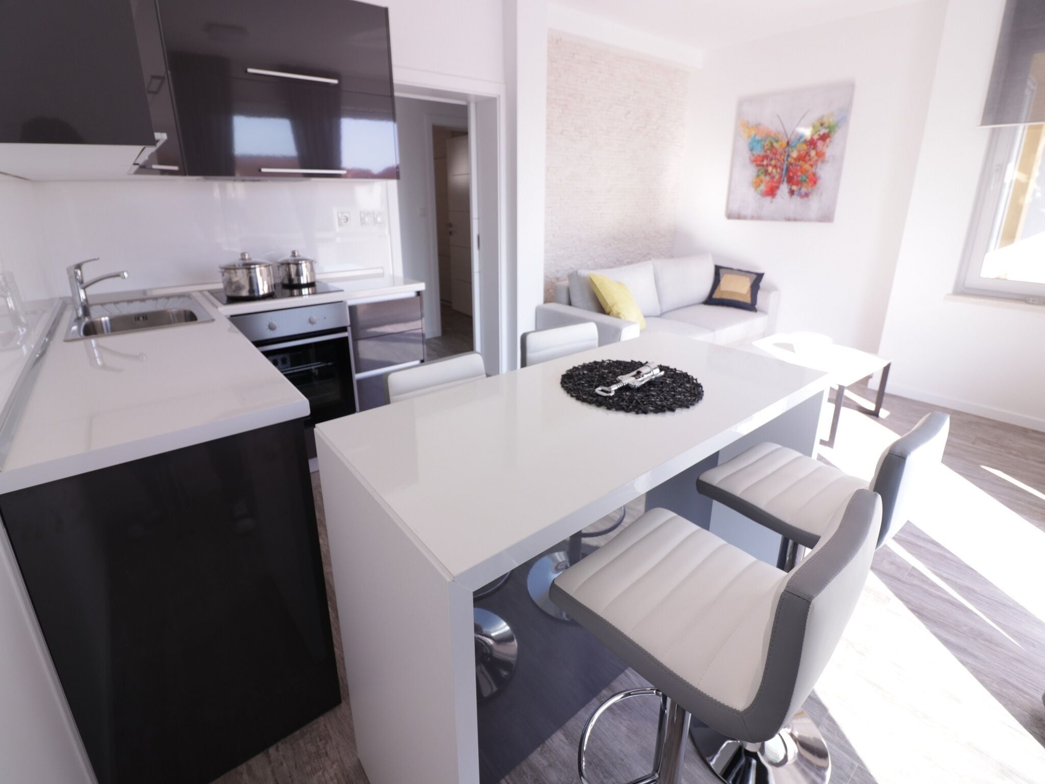 Cocina moderna con isla central, encimeras blancas y electrodomésticos de acero inoxidable. Hay un comedor con mesa y sillas blancas, y un sofá con un cojín amarillo. Se aprecia una obra de arte colorida en la pared.