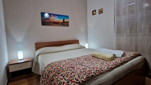 A neatly made bed with a patterned blanket, a bedside lamp, and a painting of a sunset on the wall. - View of the gulf of castellammare mountains and beach (Alcamo Marina)