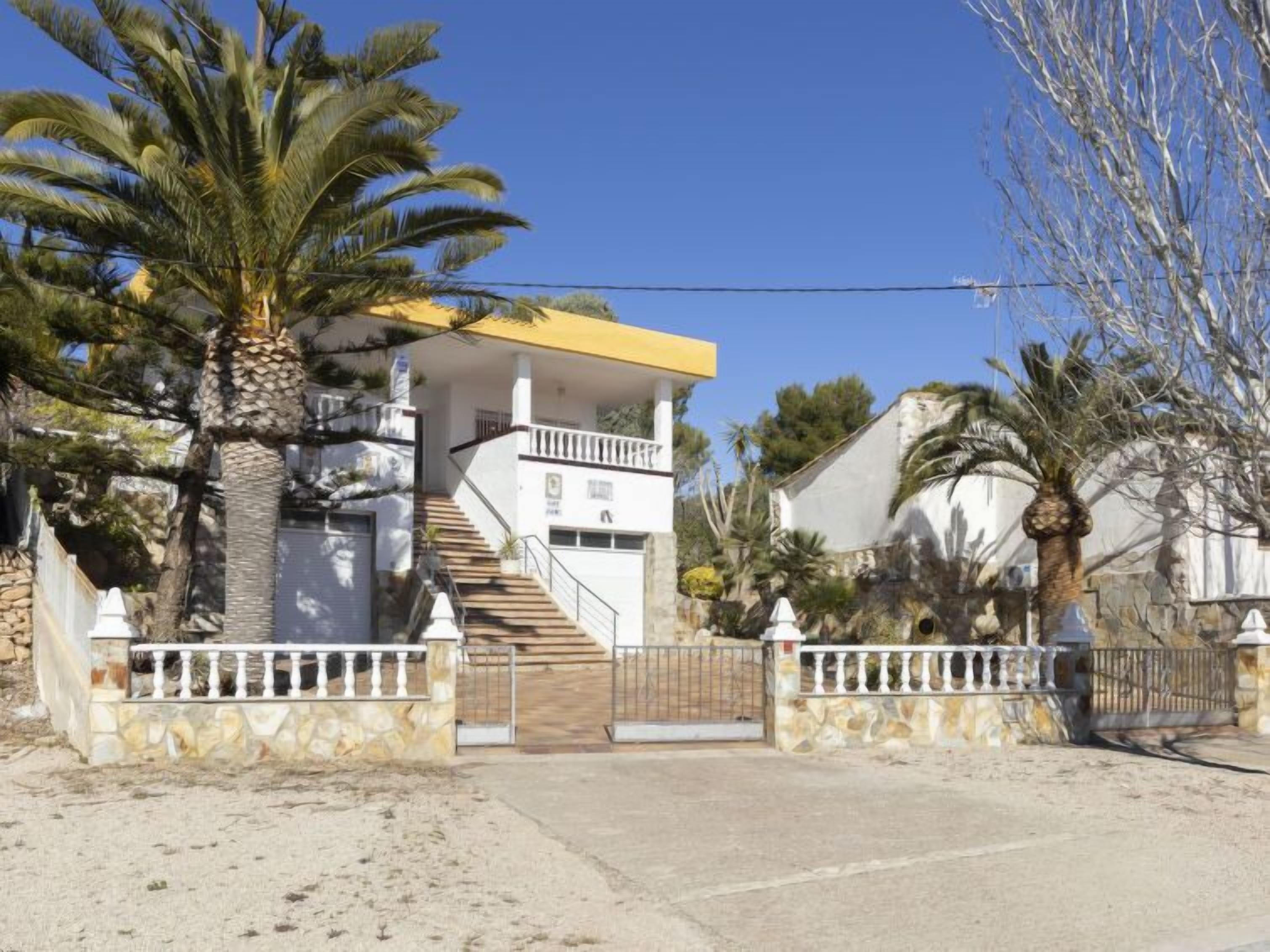 Een huis met twee verdiepingen, met een trap naar een balkon, omringd door palmbomen en een stenen muur.