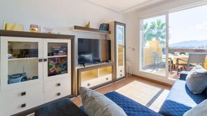 A modern living room with a TV, storage cabinets, and a large sliding glass door leading to an outdoor seating area with a view of palm trees and mountains. - Lomas del Mar V by Interhome (Vera)