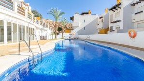 A swimming pool with clear blue water, surrounded by white buildings with balconies and a stone wall. - Lomas del Mar V by Interhome (Vera)