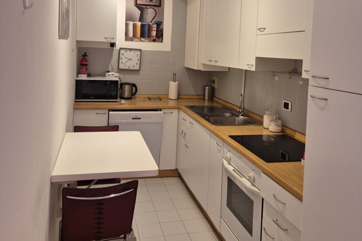 Una cocina compacta con armarios blancos, microondas, fregadero y un rincón comedor con una silla.