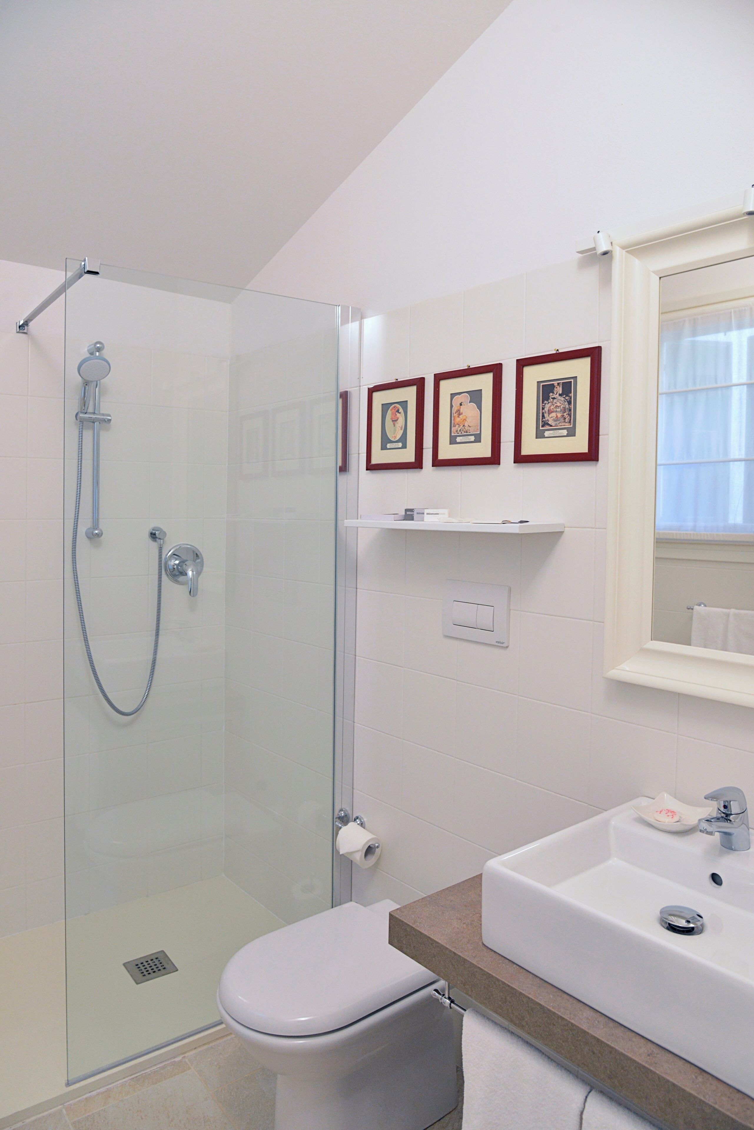 A bathroom with a glass shower enclosure, a toilet, a sink, and three framed pictures on the wall.