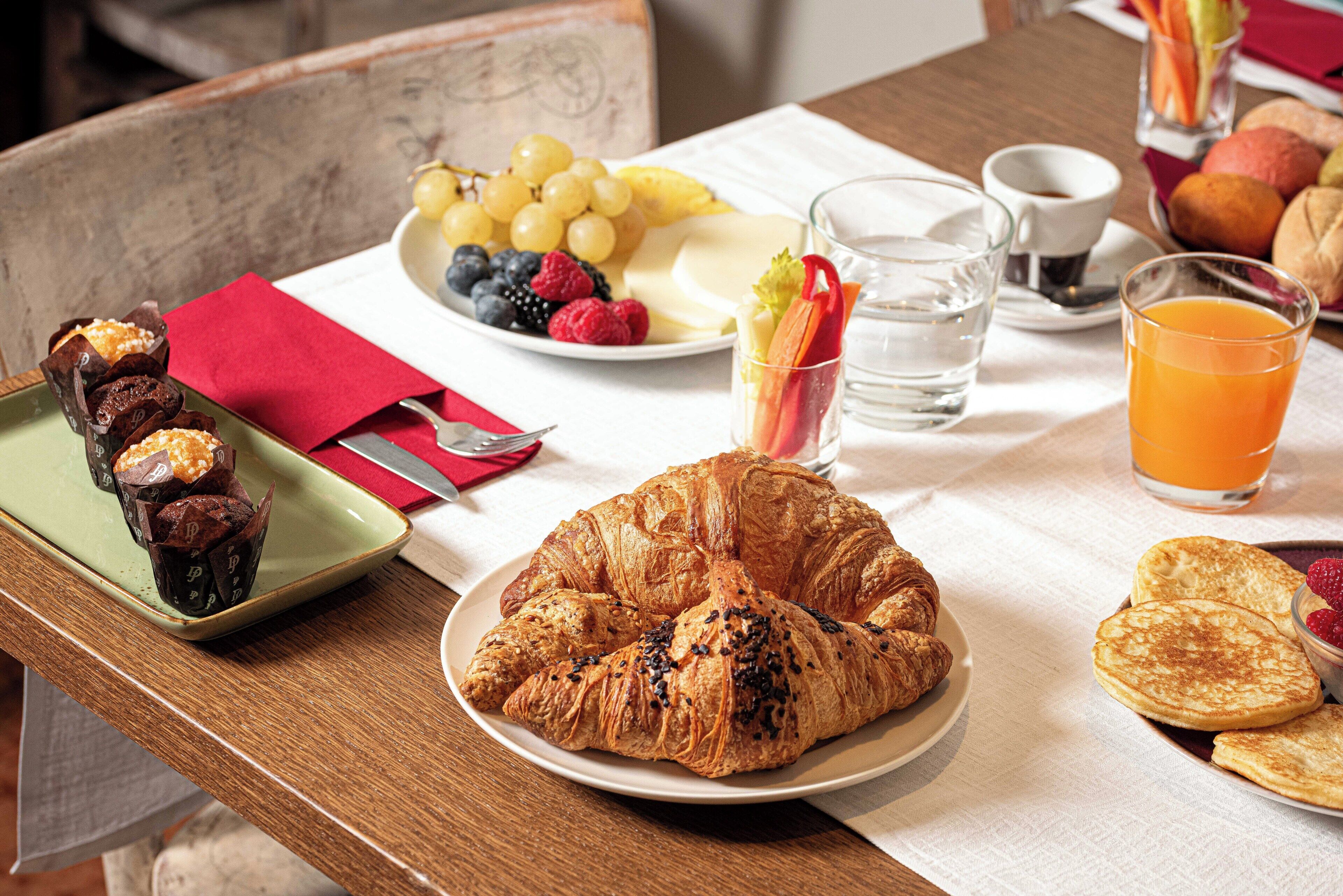 Disposição para o pequeno-almoço com croissants, frutas e bebidas sobre uma mesa.