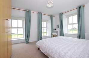 Ein Schlafzimmer mit einem großen Bett, zwei Fenstern mit Vorhängen, einem Heizkörper und Blick ins Freie.