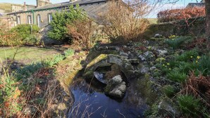 Eine Steinsbrücke über einen kleinen Bach in einem Garten mit einem Steinhaus im Hintergrund.