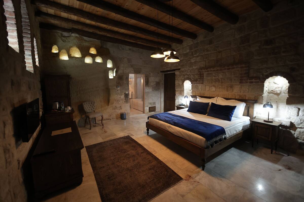 A rustic bedroom with a large bed, wooden beams, stone walls, and hanging light fixtures.