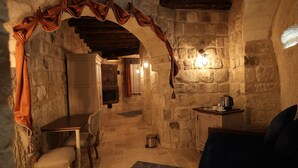 A cozy stone-walled room with a wooden ceiling, a dining table with chairs, a sofa, and a small kitchen area with a kettle.