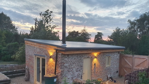 Un pequeño edificio de ladrillo con chimenea, una puerta y dos ventanas, rodeado de árboles y una valla de madera.