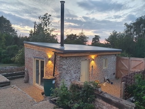 Un pequeño edificio de ladrillo con chimenea, una puerta y dos ventanas, rodeado de árboles y una valla de madera.