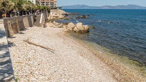 A pebble beach with clear water, a stone wall, and a staircase leading to a building. - Freya: Apartment in the center of the village (L'Escala)