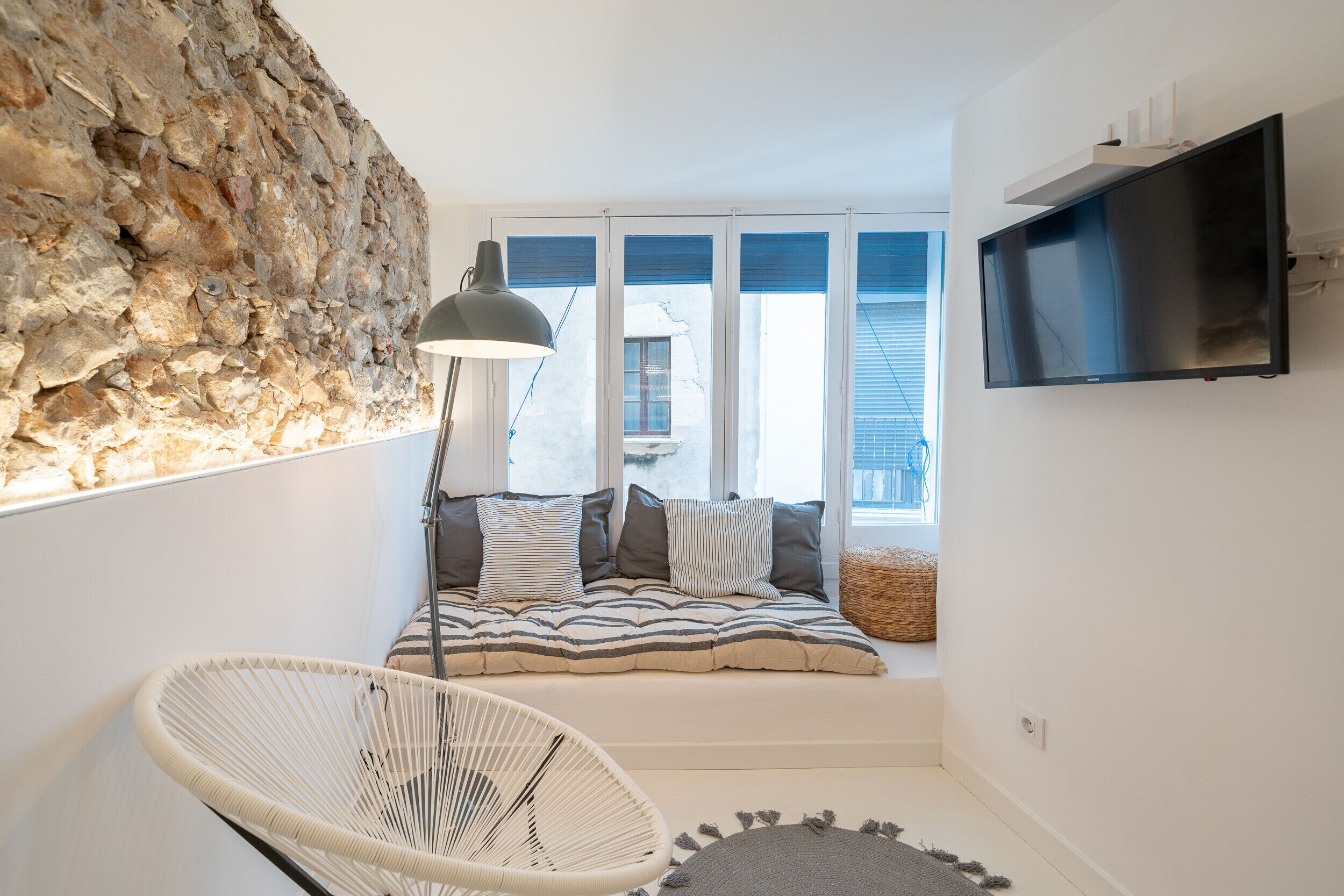 A cozy room with a stone wall, a bed with striped bedding, a white chair, and a TV on the wall.