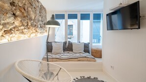 A cozy room with a stone wall, a bed with striped bedding, a white chair, and a TV on the wall. - Freya: Apartment in the center of the village (L'Escala)
