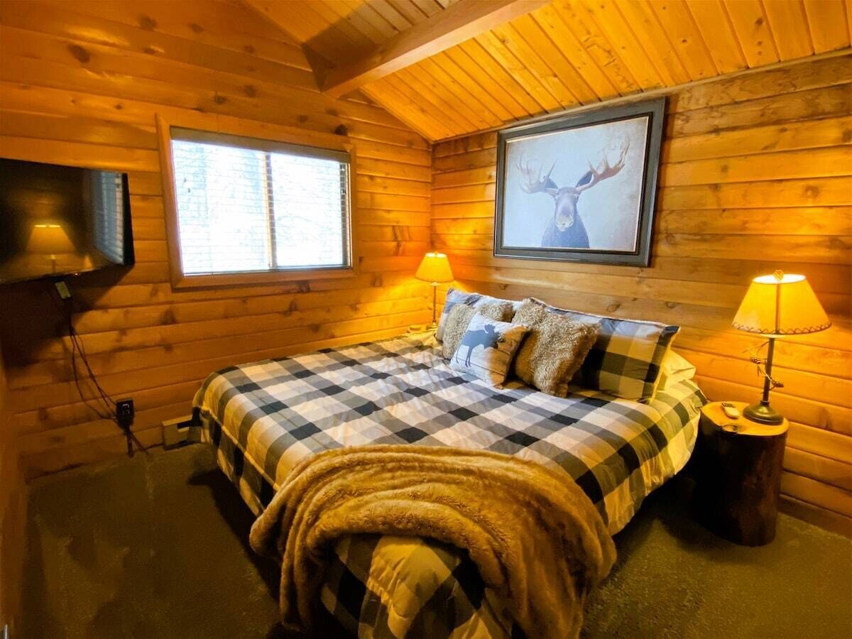Une chambre confortable dans un chalet en rondins, avec une couvre-lit à carreaux, deux lampes de chevet, un tableau représentant un cerf accroché au mur et une fenêtre avec des stores.