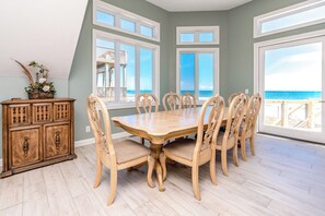 A dining room with a large table and chairs, a wooden cabinet, and large windows with a view of the beach. - @ The Beach - 7BR Oceanfront Home in Hampton Colony (North Topsail Beach)