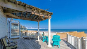 Terraza de madera con sillas y un pérgola con vistas a una playa.