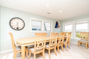 A dining area with a round table and six chairs, a wall clock, and two windows with blinds. - @ THE PEACH offers an escape to an oceanfront/oceanside (North Topsail Beach)
