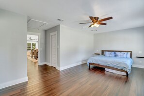 Quarto com piso de madeira, cama com cabeceira branca, ventilador de teto e porta que dá para uma sala de jantar.