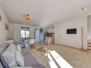 Amplia sala de estar con sofá, mesa de centro y mesa de comedor con sillas. Cuenta con una zona de cocina con refrigerador y televisor montado en la pared.
