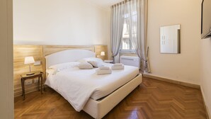 A hotel room with a large bed, wooden headboard, bedside tables with lamps, a window with curtains, and a wall-mounted air conditioning unit.