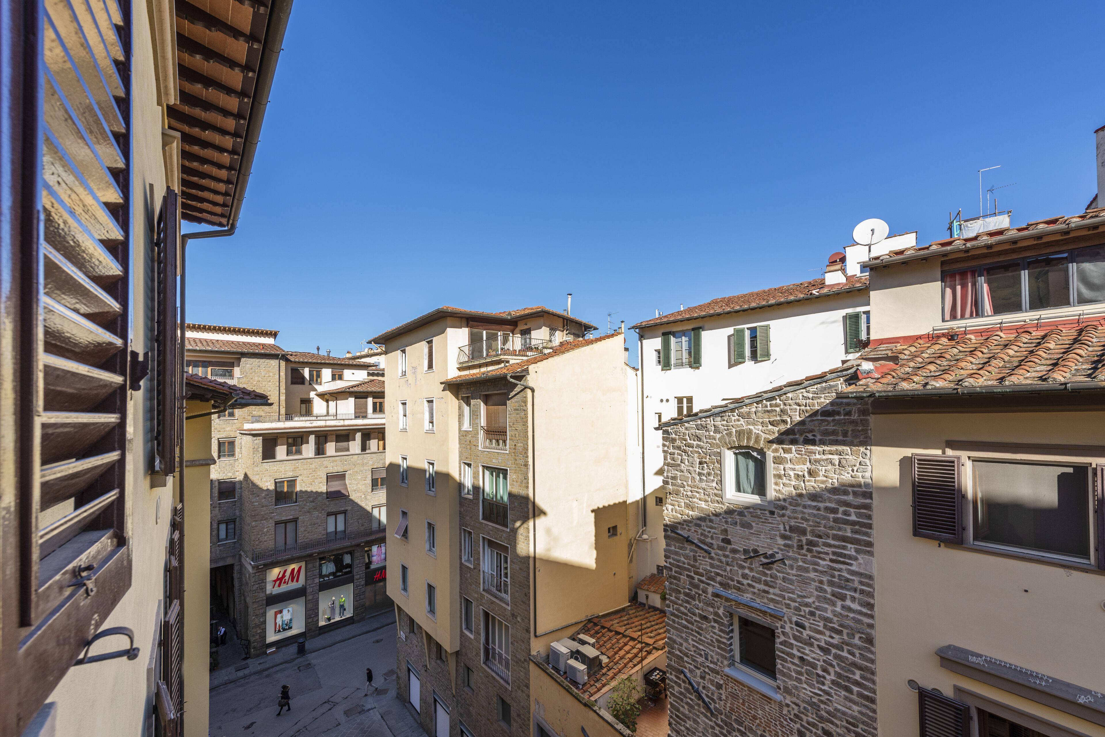 A narrow street with buildings featuring balconies and a variety of architectural styles, including stone and plaster facades.