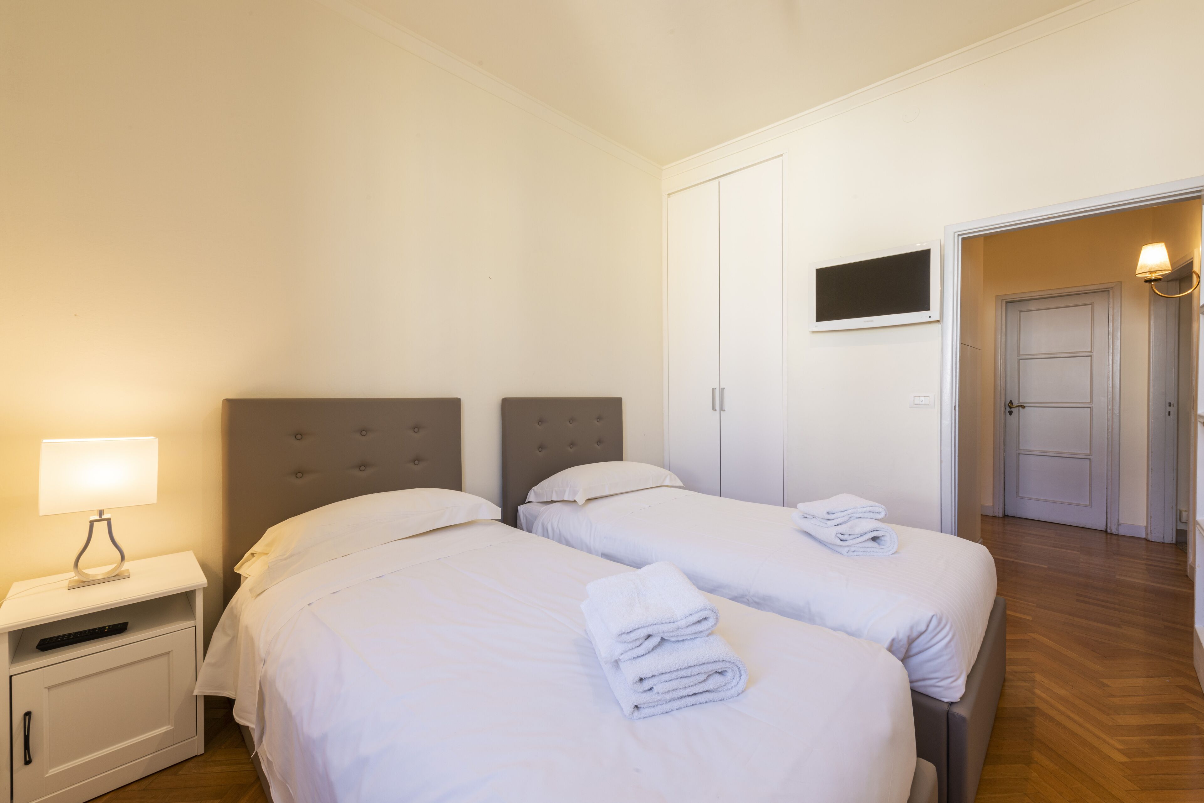Camera d'albergo con due letti, un comodino con una lampada, una TV a parete e una porta che dà su un'altra stanza.