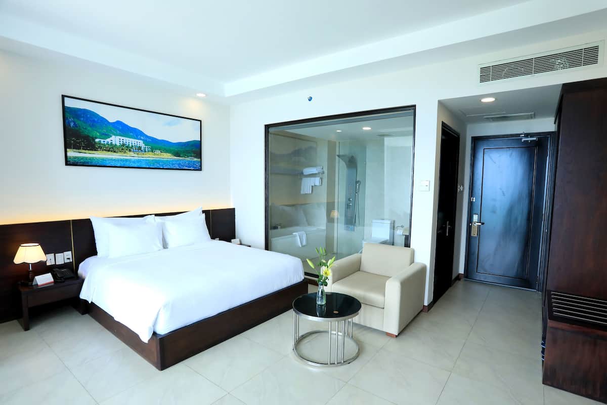 Quarto de hotel com uma cama grande, uma mesa de cabeceira com abajur, uma mesinha redonda com um vaso, um sofá e uma porta de vidro deslizante para um banheiro.