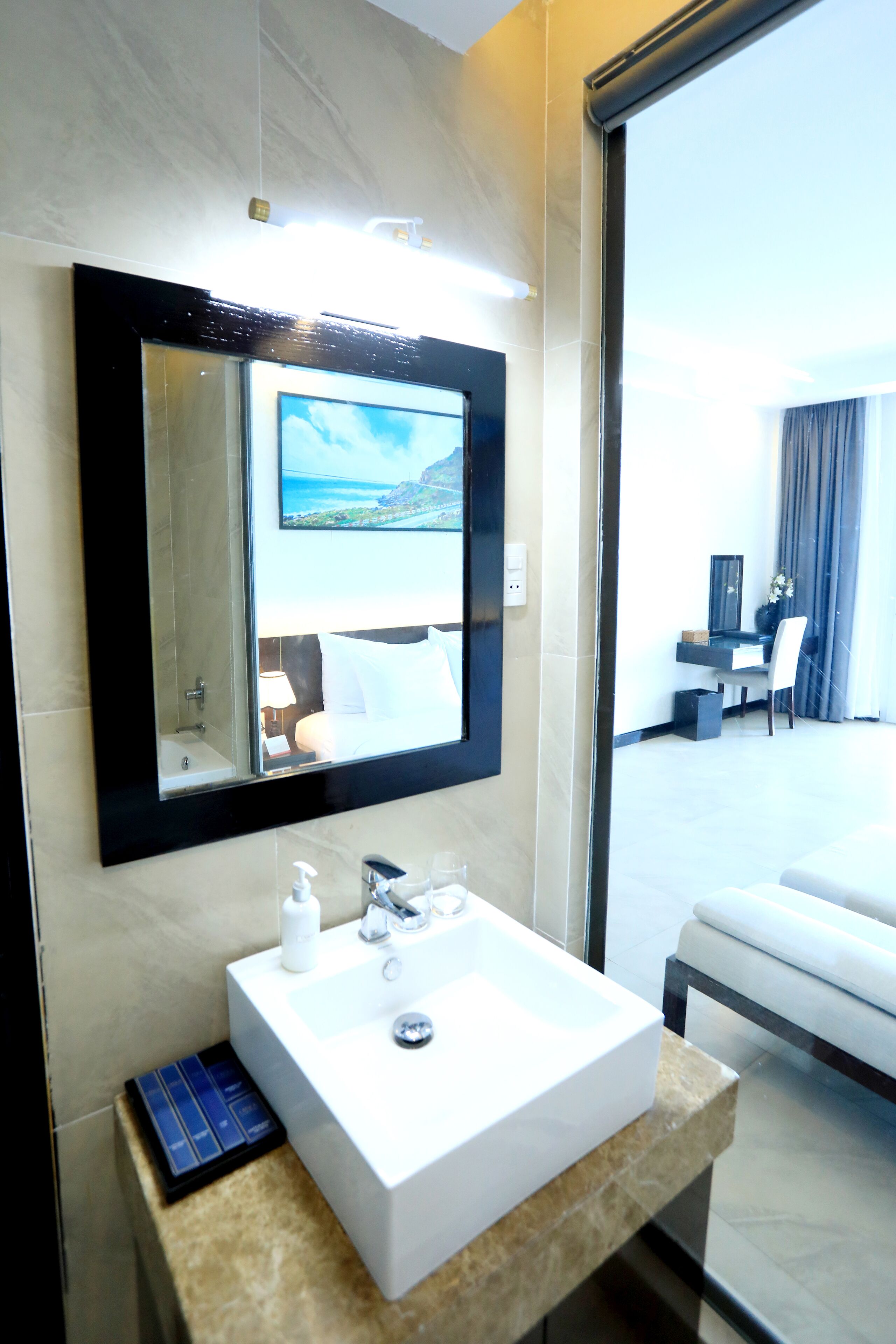 A hotel bathroom with a large mirror, a white sink, and a view of a bedroom through the glass door.
