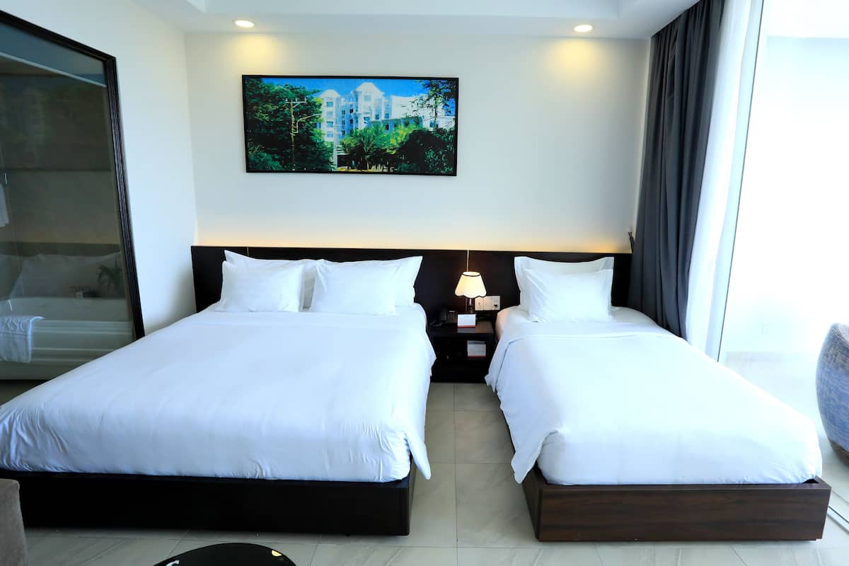 Quarto de hotel com duas camas, uma mesa de cabeceira com abajur, uma janela grande com cortinas e um quadro na parede.