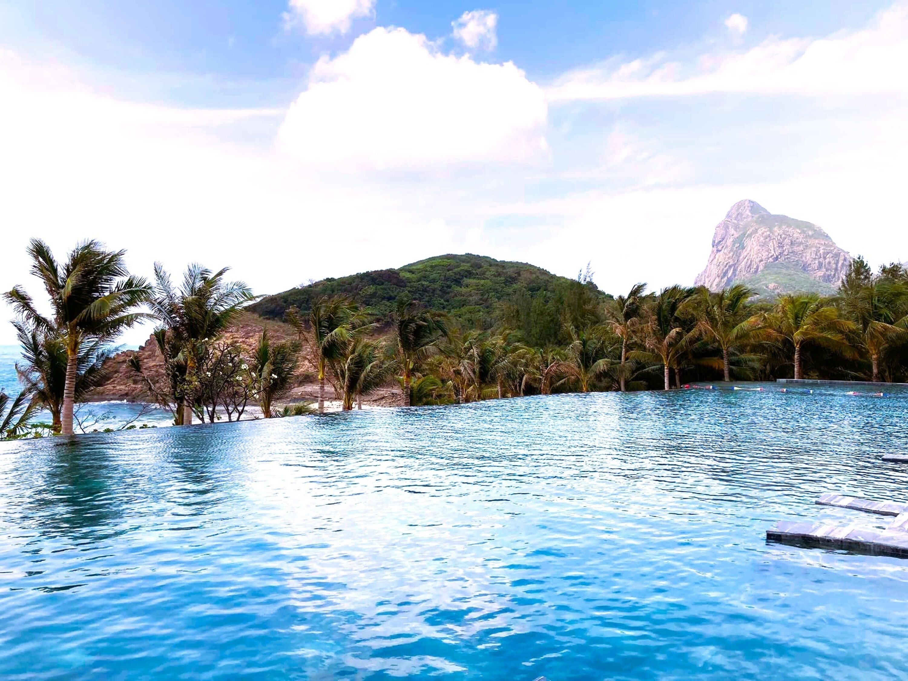 Una piscina azul cristalina rodeada de palmeras y con una montaña al fondo.