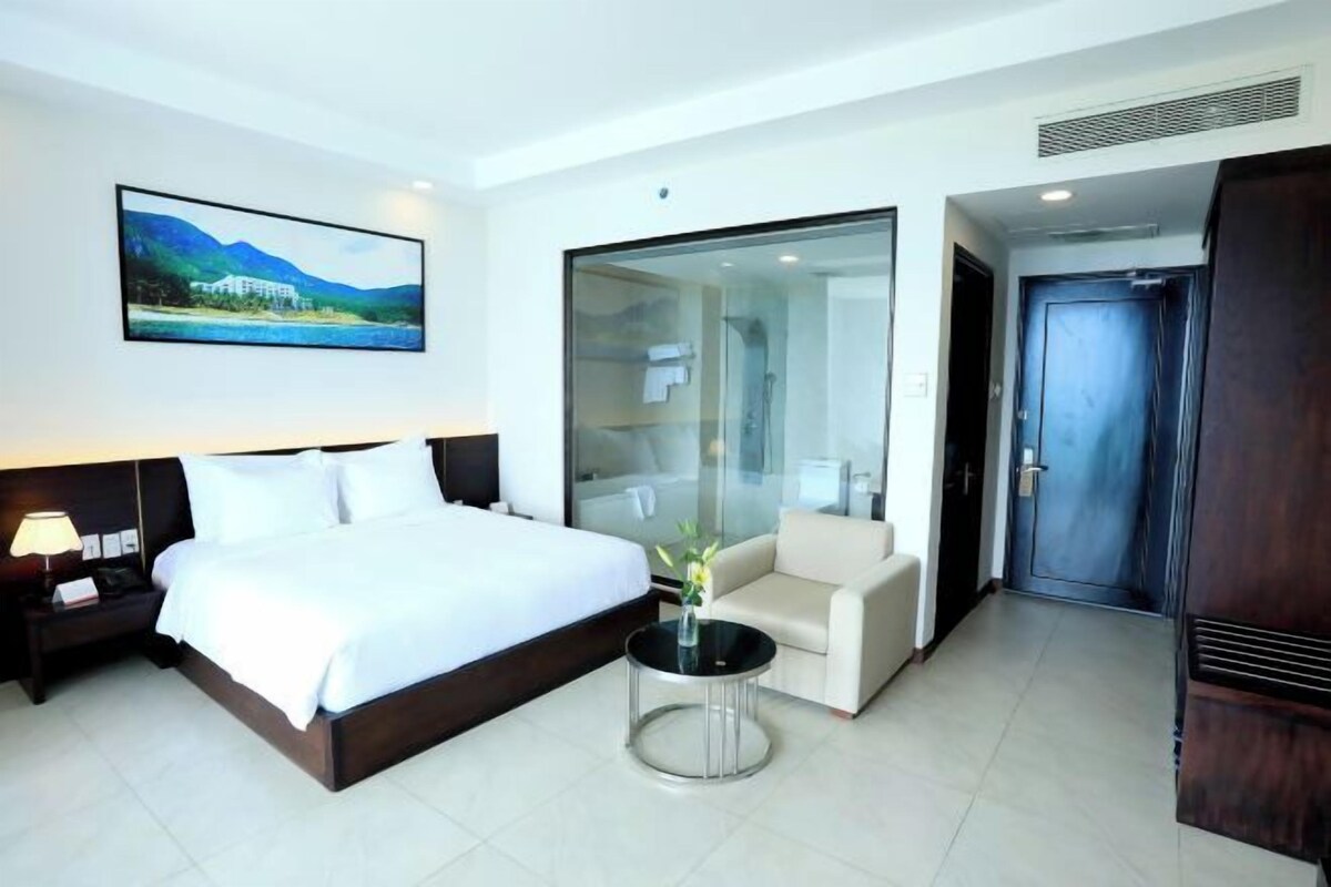 A hotel room with a bed, a chair, a small table, a painting on the wall, and a door.