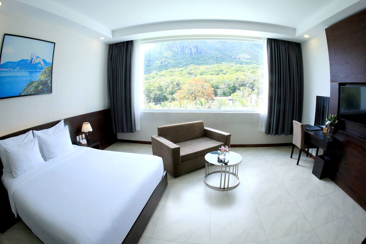 Quarto de hotel com uma cama grande, um sofá, uma mesinha redonda com um vaso, uma escrivaninha com uma TV e uma janela com cortinas.