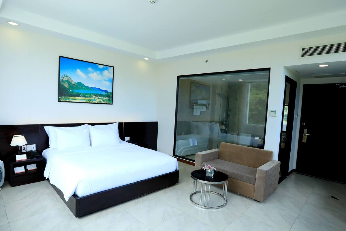 Quarto de hotel moderno com uma cama grande, um sofá, uma mesa pequena e um box de chuveiro de vidro.