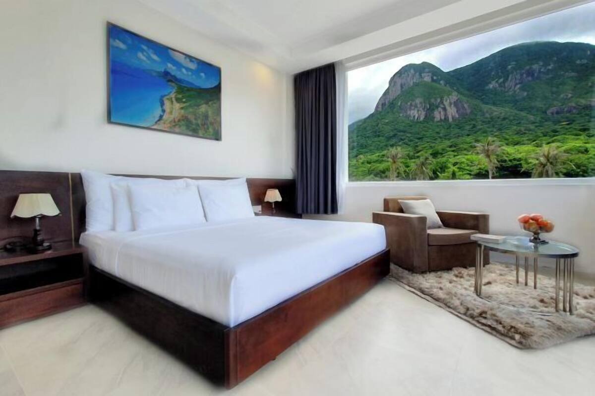 Quarto de hotel com cama grande, escrivaninha, cadeira, mesa pequena e vista para montanhas e vegetação.