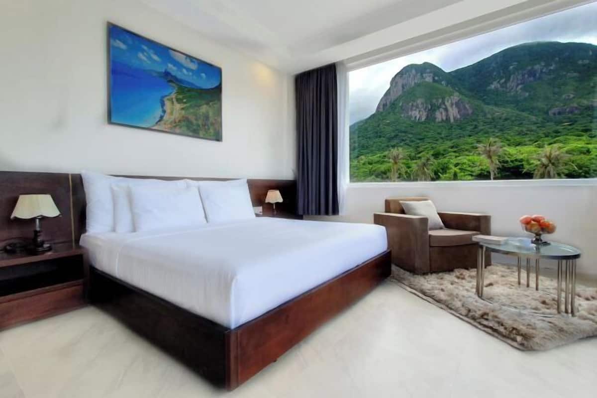 A hotel room with a large bed, a desk, a chair, a small table, and a view of mountains and greenery.