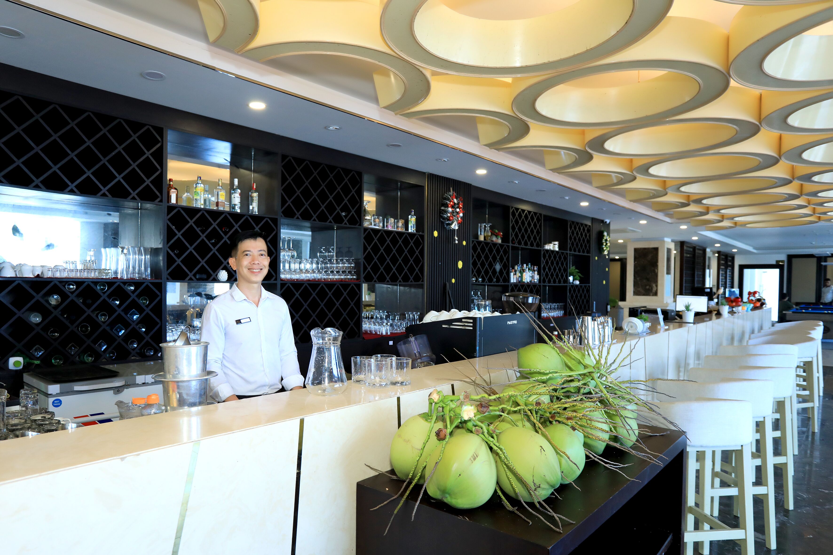 A bar area with a bartender, shelves with bottles, and a decorative plant arrangement.