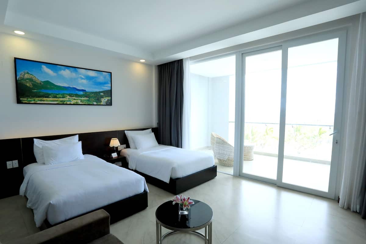 Quarto de hotel com duas camas, uma janela grande, um quadro na parede e uma mesinha com um vaso.