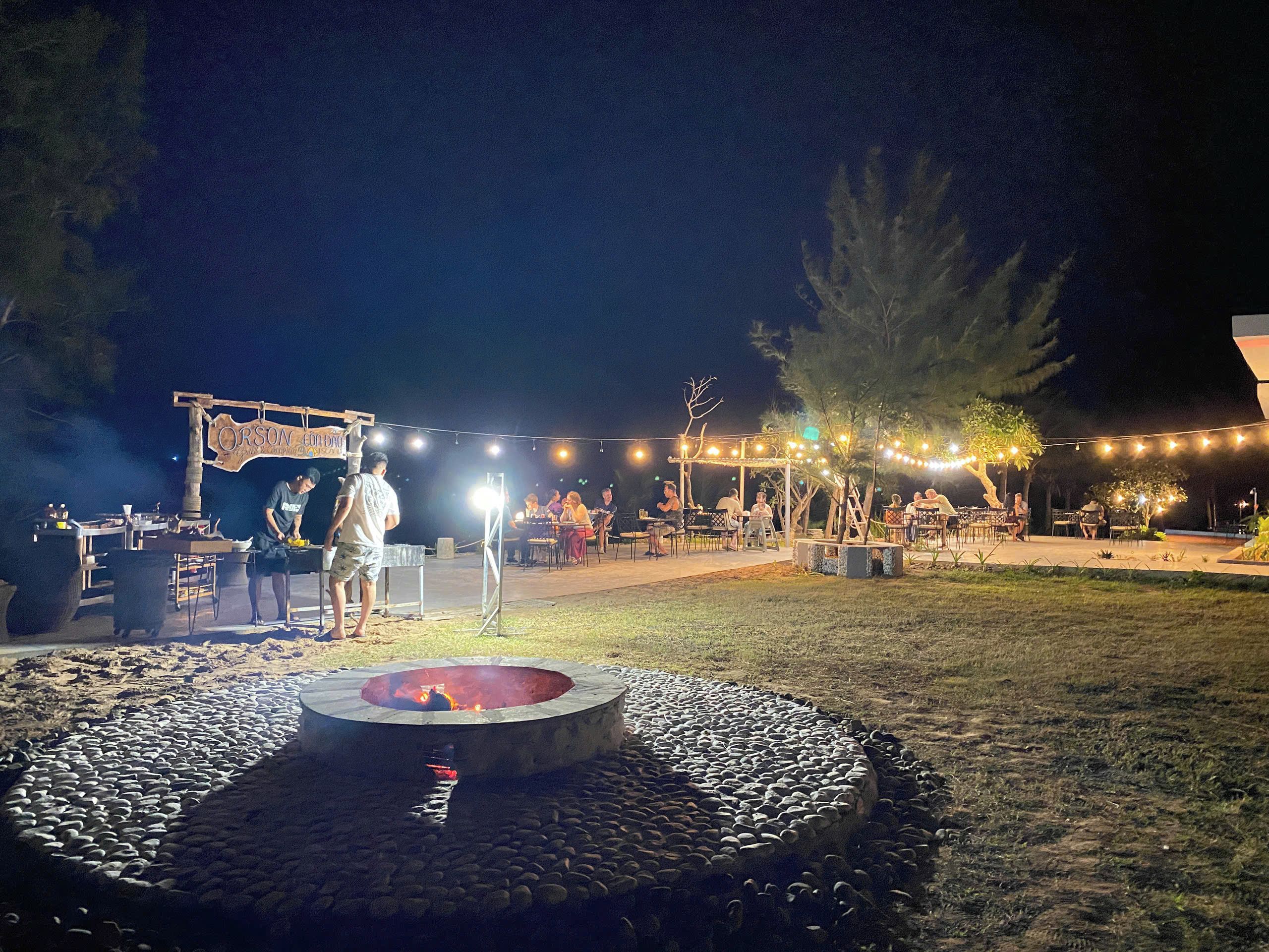 Evento al aire libre con un brasero rodeado de piedras, mesas con gente y luces colgantes.