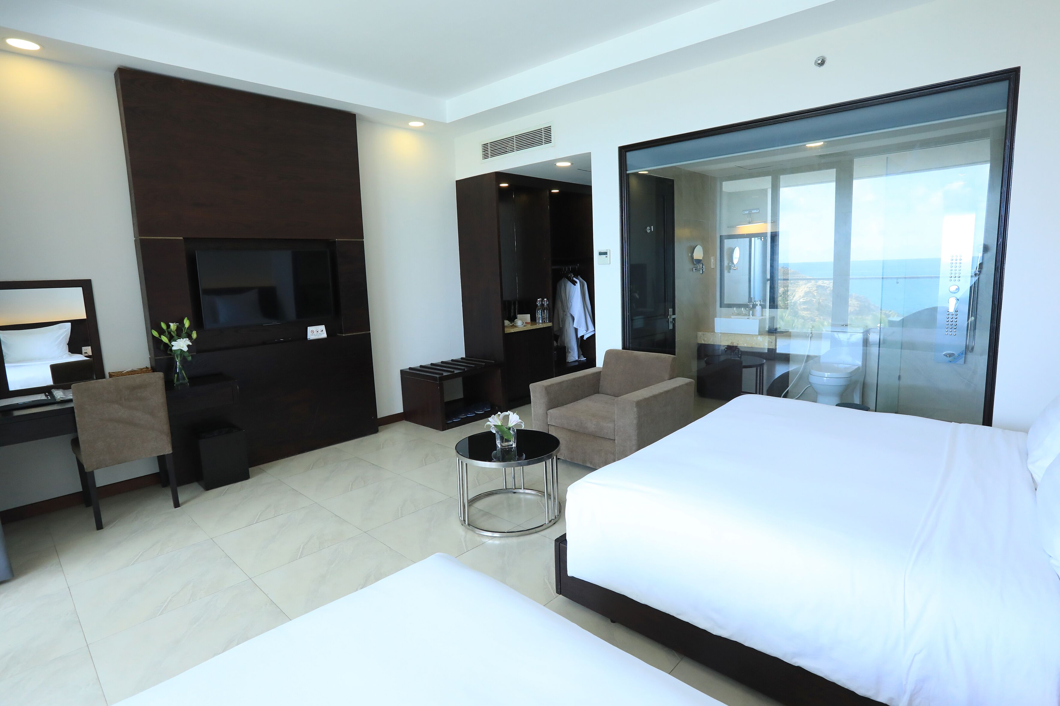 A modern hotel room with a large bed, a desk with a computer, a TV, a seating area with a chair and a small table, and a view of the ocean through a glass door.