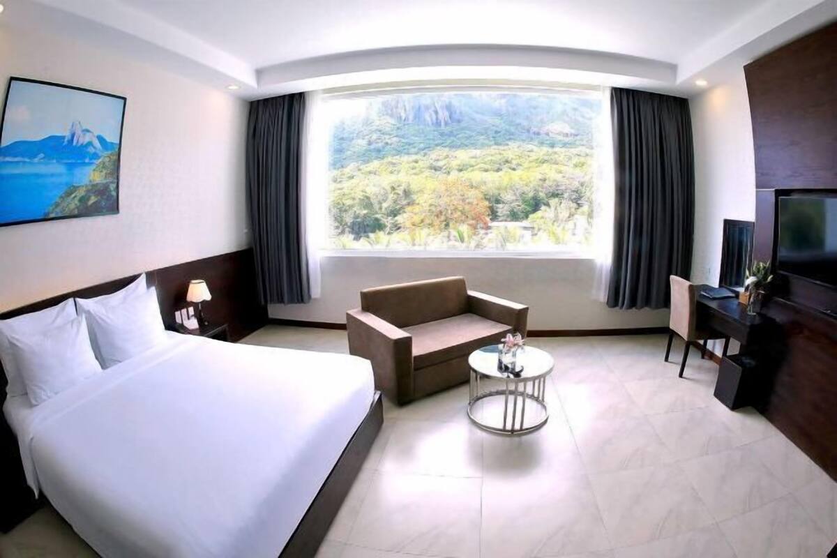 Quarto de hotel com cama, sofá, mesa redonda pequena, televisão e uma grande janela com vista para uma paisagem montanhosa.