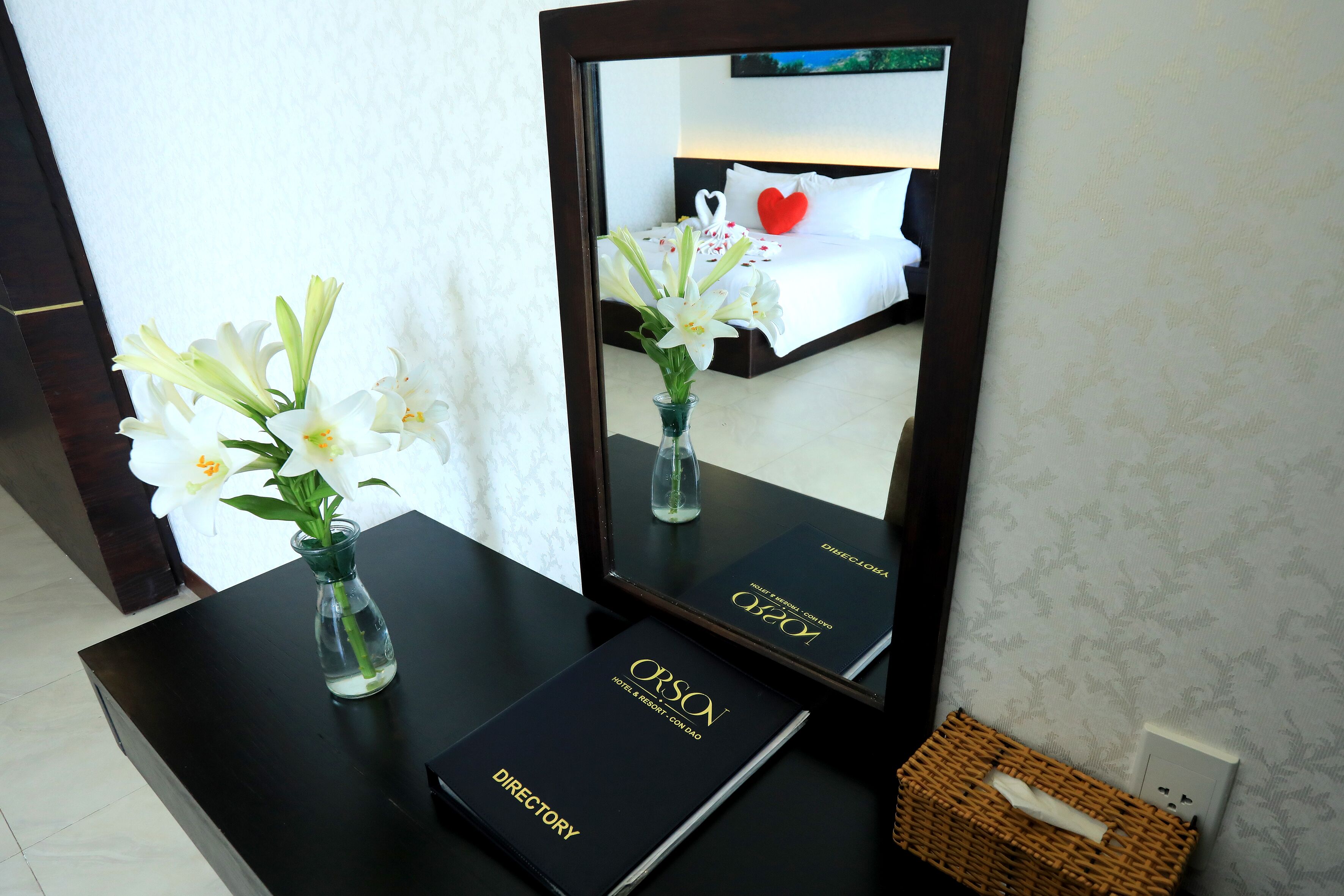 A hotel room with a bed, a vase with flowers, a mirror, and a directory on a table.